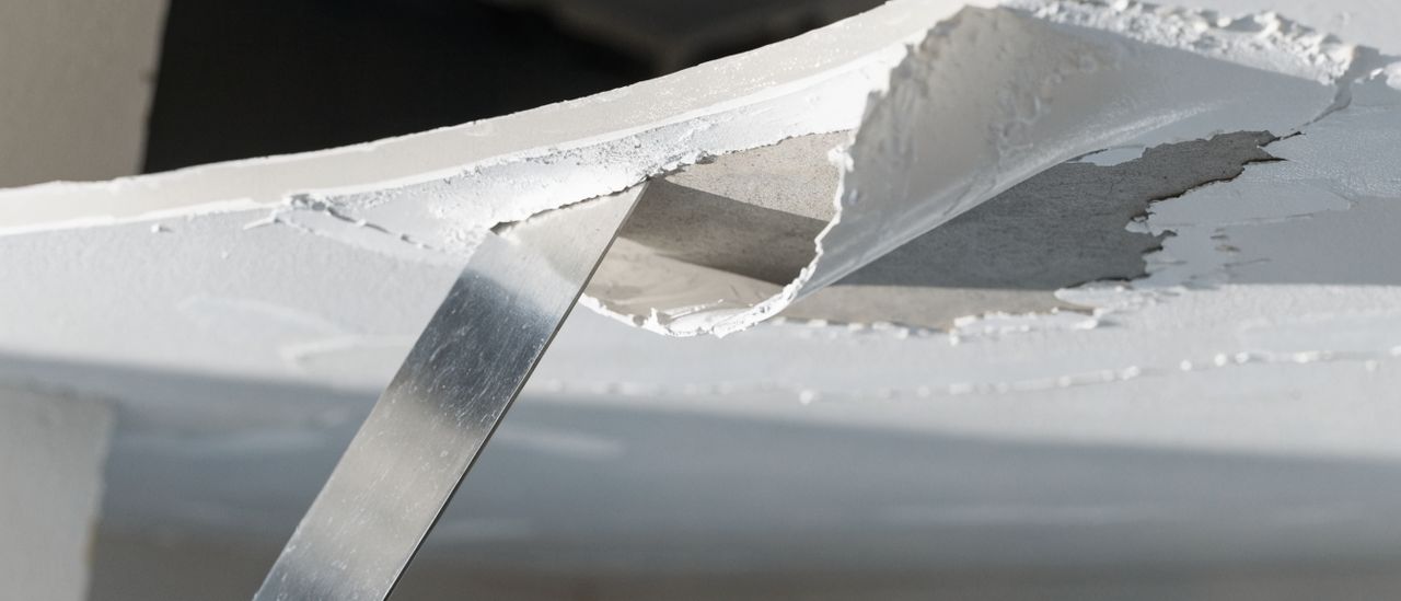 Close-up of a scraper removing wet popcorn ceiling texture from drywall